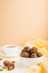 Chocolate caramel ball candies with almonds and a cup of coffee on a white and orange background. Side view, copy space.