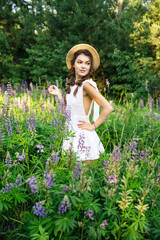 a young beautiful girl in a white dress and hat walks across the field with lupins