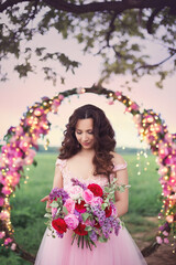 Beautiful bride in pink dress standing with her bouquet at a round flower arch under a large single tree in a field at sunset. A destination summer wedding, romantic country ceremony.
