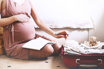 Pregnant woman packing bag for maternity hospital, making notes, checking list in diary. Expectant...