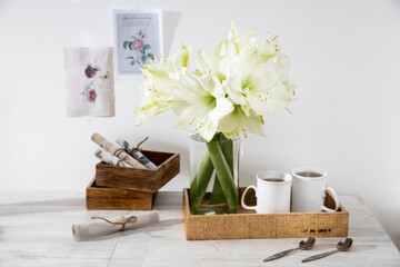 A bouquet of white lily in a glass vase on a table with two tall cups of coffee, a teapot, spoons, and a milk jug. Copy space