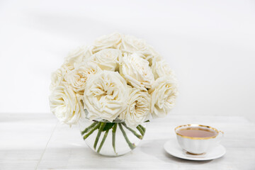 A bouquet of white roses in a glass vase on a table with cups of tea on the table.