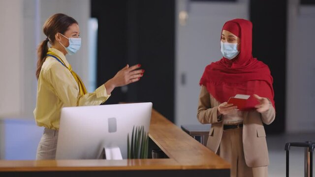 Polite caucasian young woman flight assistant servicing Muslim hijab arabic tourist checking passport visa at registration counter. Check-in. Airport.