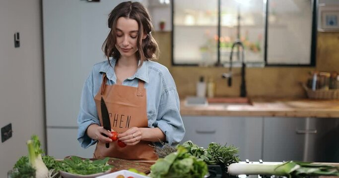 Young cheerful housewife making salad on a kitchen table full of healthy green food ingredients. Vegan organic food, healthy cooking concept
