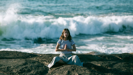 A young yoga woman meditates in the lotus position sitting on rocks on the Alantic ocean coast.