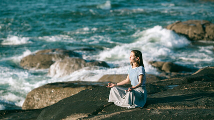 Young yoga woman meditates in the lotus position sitting on rocks on the Alantic ocean coast.