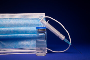 Vaccine bottle phial with no label near blue medical mask and medical syringe with injection...