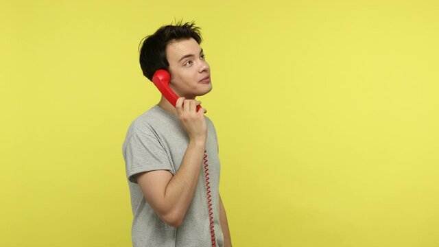 Profile portrait bored young guy holding red handset, talking wired landline phone with relatives, listening carefully and answering questions. Indoor studio shot isolated on yellow background