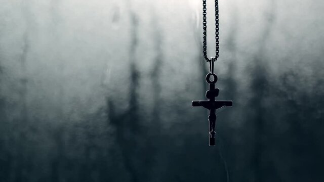 Christian crucifix pendant swinging on chain in front of rain drenched window