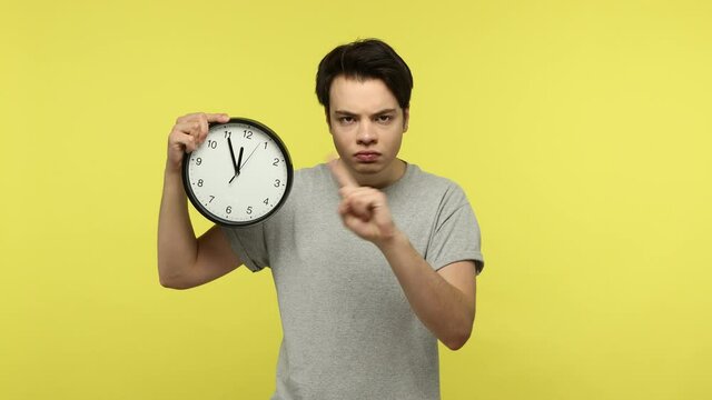 Bossy strict teenager in gray t-shirt pointing finger at big wall clock in his hand and scolding you for being late, time management. Indoor studio shot isolated on yellow background