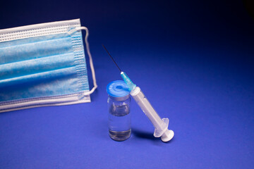 Vaccine bottle phial with no label near blue medical mask and medical syringe with injection...