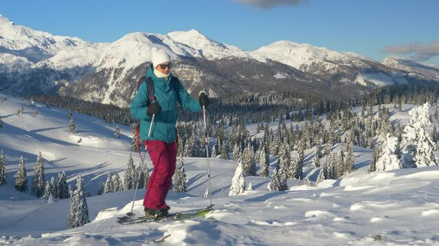 CLOSE UP: Young female tourist on splitboarding trip hikes up a snowy hill. Fit Caucasian woman ski touring in Velika Planina on a perfect sunny winter day. Splitboarder ascends up a scenic mountain.