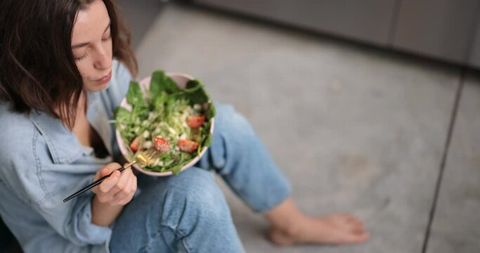 Young woman eating green salad while sitting on the kitchen floor. Healthy lifestyle concept. High angle dolly out shot