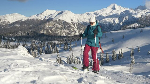 CLOSE UP: Fit Caucasian woman ski touring in Velika Planina on a perfect sunny winter day. Young female tourist on splitboarding trip hikes up a snowy hill. Splitboarder ascends up a scenic mountain.