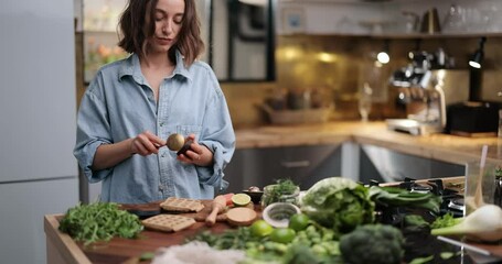 Young woman making healthy breakfast, cutting avocado on the kitchen table full of greens and vegetables at home. Healthy eating and lifestyle concept - Powered by Adobe