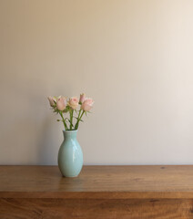 Vertical closeup of pale pink roses in green vase on oak table against beige wall with copy space
