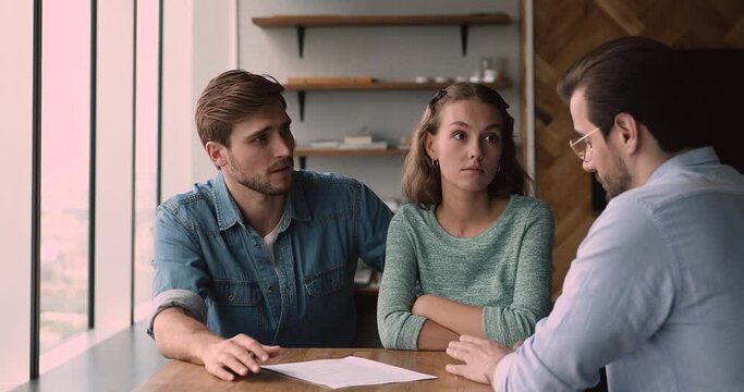 Confused young family couple dissatisfied with contract agreement terms of conditions change, arguing with agent at office meeting. Unhappy clients blaming financial advisor, demanding compensation.