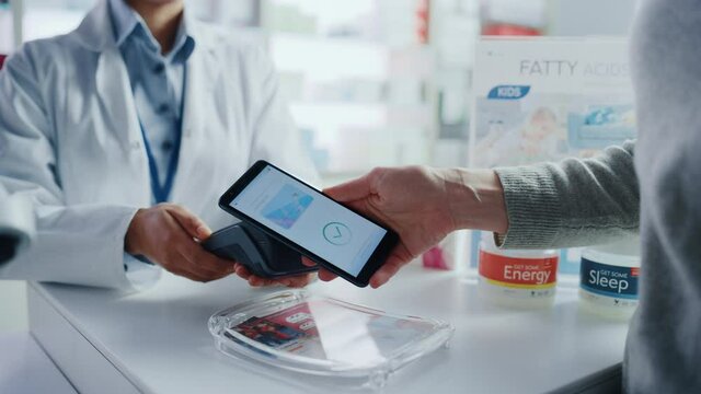 Pharmacy Drugstore Checkout Cashier Counter: Pharmacist and a Customer Using NFC Smartphone with Contactless Payment Terminal to Buy Prescription Medicine, Health Care Goods. Hand Focus Closeup