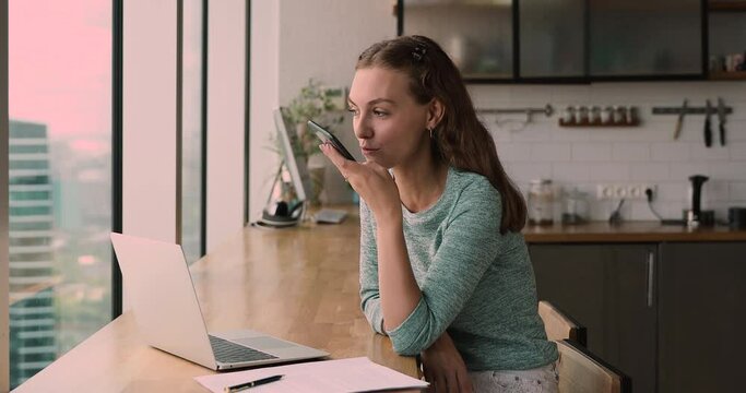 Happy millennial business lady recording audio message, using voicemail, activating artificial intelligence assistant mobile app, web surfing or translating information, working distantly at home.
