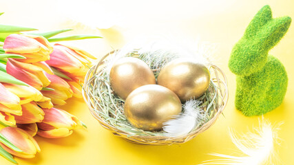 Easter eggs Golden color in basket with spring tulips, white feathers on pastel yellow background in Happy Easter decoration. Festive decoration.