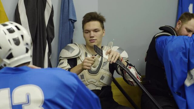 Tilt up shot of young handsome sportsman sitting in locker room, taping stick and talking with teammates before hockey game