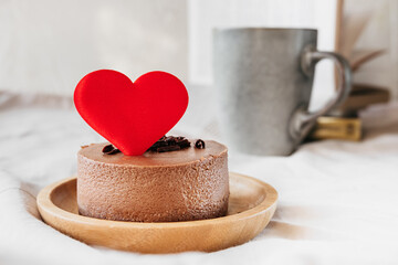 red heart decoration of round small chocolate cheesecake without baking on bamboo plate, cup with drink and book are blurred on background. cute surprises and gifts for valentines day, selective focus
