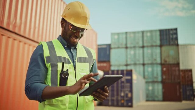 Smiling Portrait of a Handsome African American Black Industrial Engineer in Yellow Hard Hat and Safety Vest Working on Tablet Computer. Foreman or Supervisor in Container Terminal.