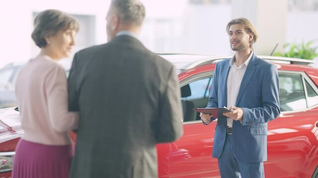 Young handsome car dealer advertising automobile to rich Caucasian senior couple choosing vehicle in dealership. Portrait of professional employee selling car to clients in showroom.