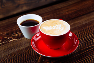 Aromatic coffee in a red cup with milk foam and latte art and freshly brewed coffee in a white cup on a dark wooden table. The choice of coffee.