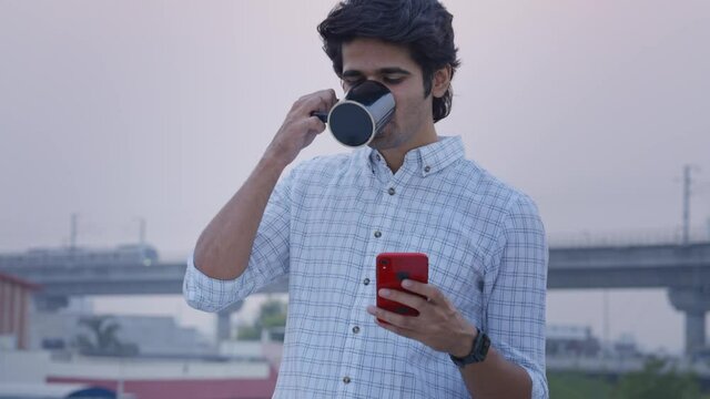 shot of a young indian male entrepreneur standing outdoors using a mobile phone while sipping a coffee from a mug with a blurry background of city metro train passing by