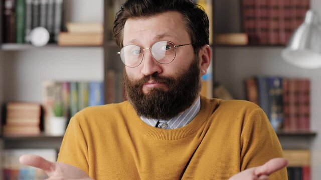 Shrug, i dont know. Perplexed bearded man in glasses in office or apartment room looking at camera and emotionally squeezes his shoulders and spreads his hands to show his ignorance. Close up