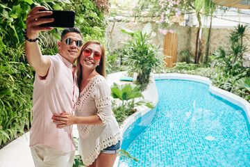 Happy Asian couple in sunglasses hugging and taking selfie by swimming pool at spa resort