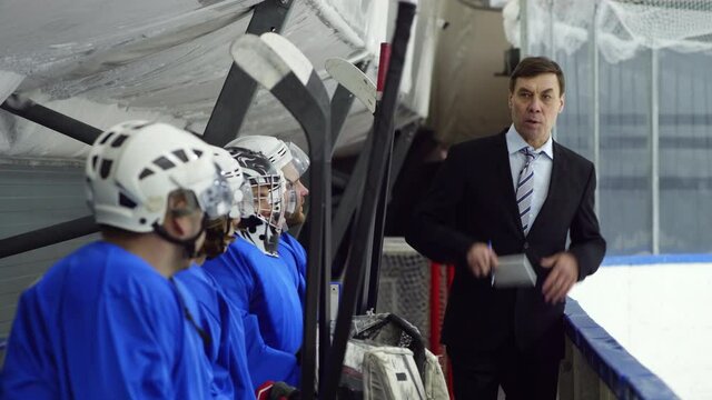 Male coach in formal suit holding notepad and pen, walking along bench on ice rink and explaining strategy to team of hockey players