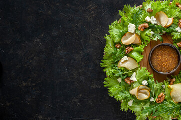 Light diet salad with pear, nuts and blue cheese. The salad is laid in the form of a wreath on a round wooden board with sauce in the center on a dark background. Copy space Top view