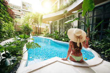 Beautiful woman in big straw hat sitting on edge of swimming pool and looking at beautiful water and lush plants around, view from the back