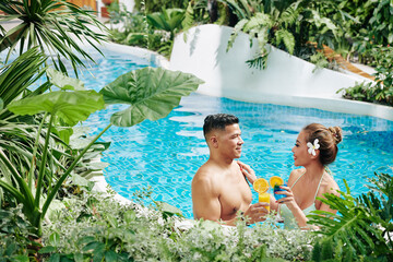 Cheerful Asian boyfriend and girlfriend standing in swimming pool among lush plants and flowers,...