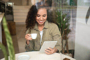 Beautiful woman with tablet computer and cup of hot espresso in cafe, view through glass window