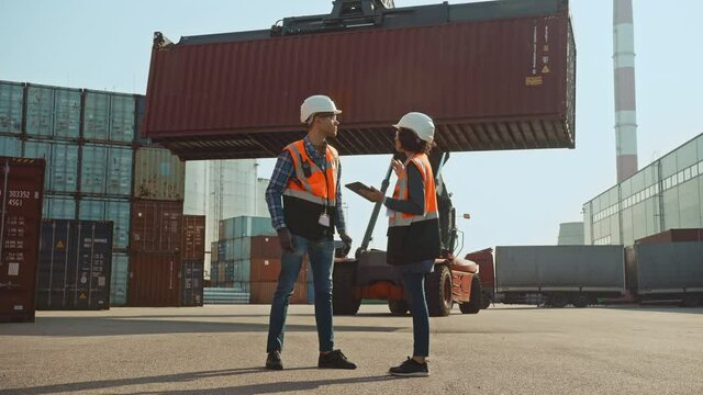 Multiethnic Female Industrial Engineer with Tablet Computer and Male Foreman Worker in Hard Hats and Orange High-Visibility Vests Stand in Container Terminal. Colleagues Talk About Logistics Business.