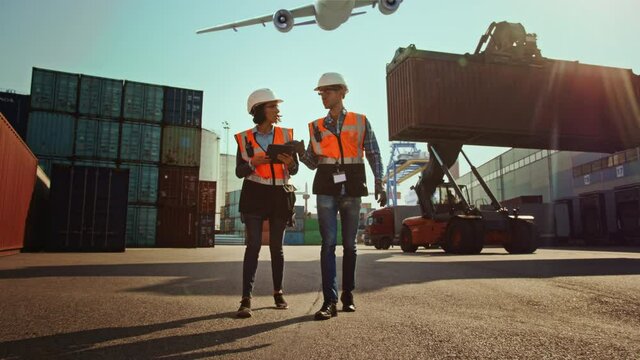 Female Industrial Engineer with Tablet Computer and Male Foreman Worker in Hard Hats Walk in Container Terminal. VFX Airplane Fly in the Sky and Double Girder Gantry Cranes Work in the Background.