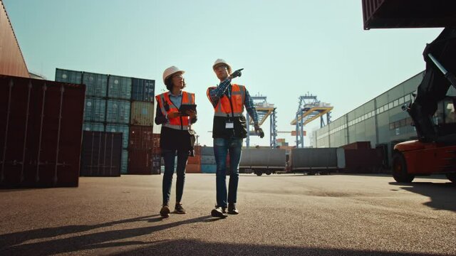 Multiethnic Female Industrial Engineer with Tablet Computer and Male Foreman Worker in Hard Hats and Safety Vests Walk in Container Terminal. VFX Double Girder Gantry Cranes Work in the Background.