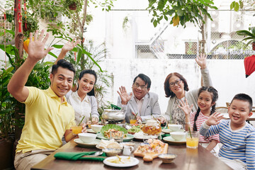 Happy big Vietnamese family sitting at big dinner with many tasty dishes table and waving with hands