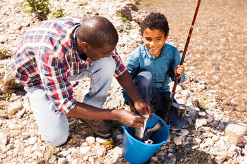 Portrait of cheerful afro man and his son holding rod and bucket of fish catch