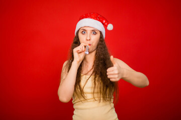 Young woman in santa hat blowing festive pipe isolated on red background.