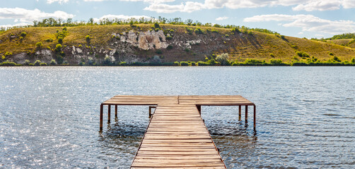 Panoramic view of wooden fishing platform on bank of sunny river with rocky hills on distance...