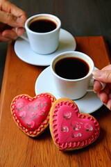 Hands of woman and man holding cup of coffee with a pair of heart shaped cookies in foreground