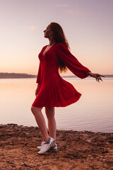 Young ginger woman in red dress at sunset. River or lake on background, pink sky. Dress in flying.