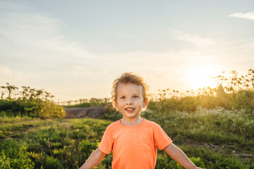 Naklejka na ściany i meble Little curly boy in orange t-shirt smile and stay in a meadow, sunset light. Boy looks up. Beautiful landscape in summer.