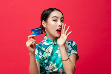 Cute Asian woman wearing oriental qipao dress showing credit card cupping hand and saying  on red studio background for Chinese new year cashless shopping concepts