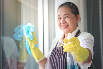 Happy smiling Asian senior elderly woman housewife wearing rubber gloves and holding cleaning spray and clothe for wiping window, spraying toward camera, cleaning up house, grandma doing housework and