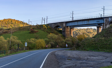 An old stone arched railway bridge over a road in the mountainous part of the Ukrainian Carpathians. Autumn mountain landscape - yellowed and reddened autumn trees combined with green needles.